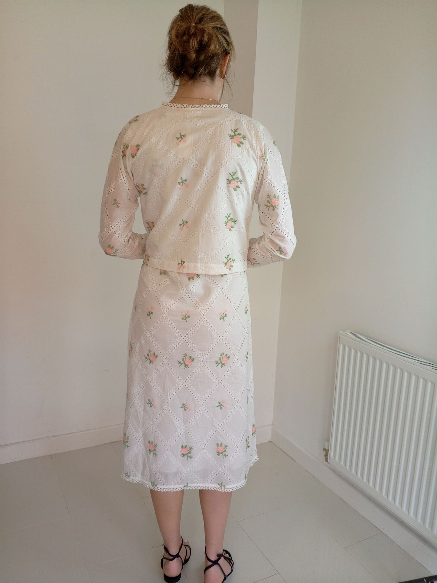 Person wearing a floral dress and cardigan in a minimal room.Cropped pink embroidered cardigan-Relaxed body fit-Round neck -Inside hidden hook closure-full lined body with100% soft fine cotton-Long sleeves with lace -Fabric: 100% Cotton-backview