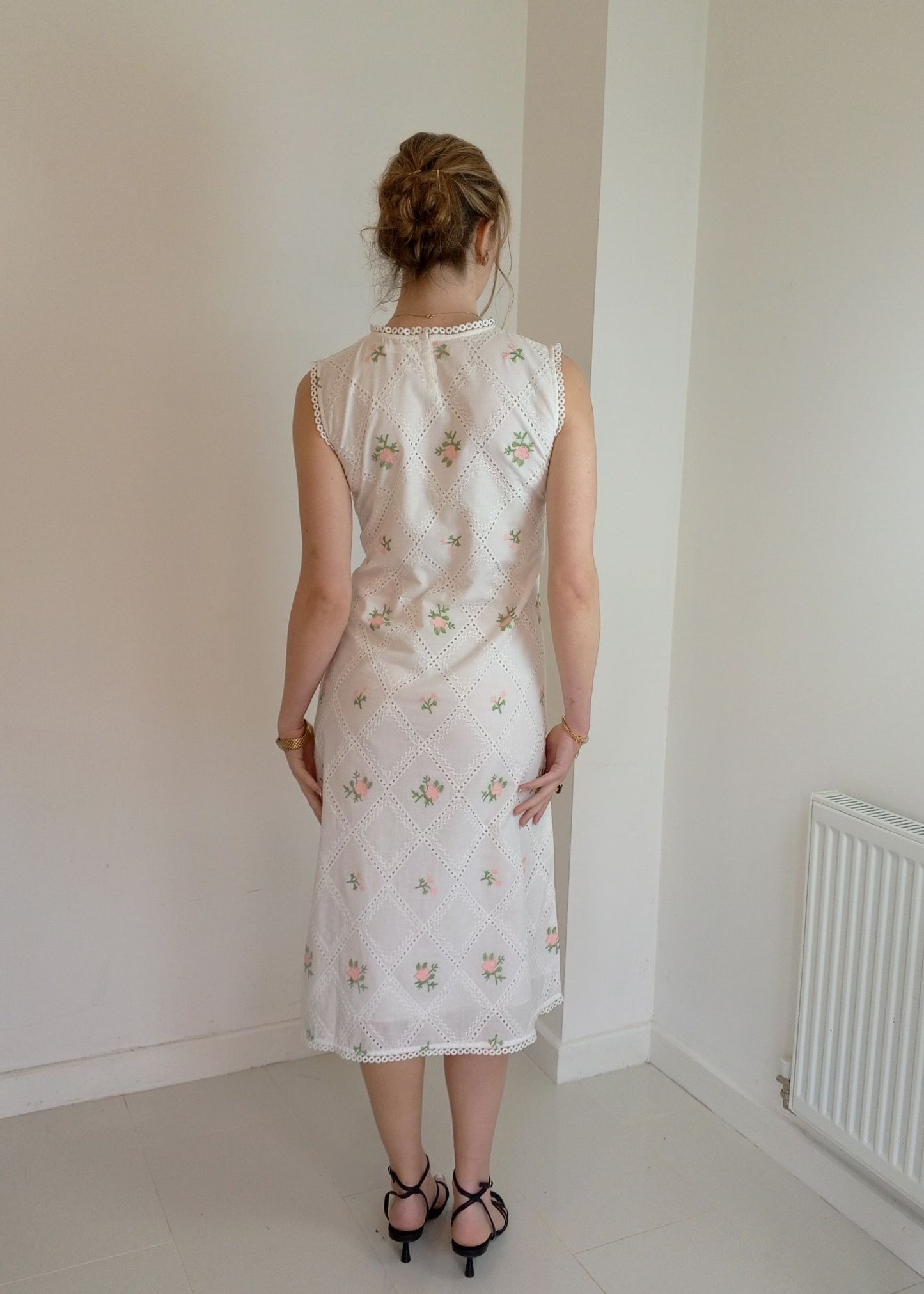 Woman wearing a white embroidered dress standing in a room Pink and white embroidered dress-Midi length relaxed body fit-Boat neck with lace -left side leg split-full lined dress with soft 100% fine cotton -Sleeveless with lace -Fabric: 100% Cotton-back angle