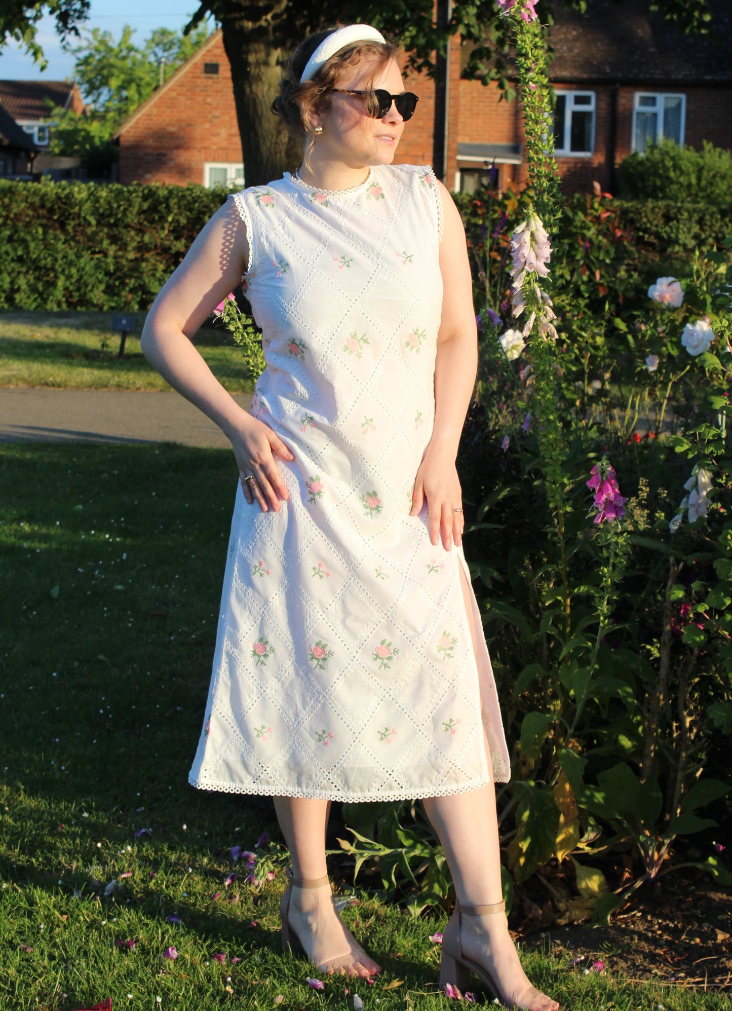 A woman standing in a garden wearing a midi-length sleeveless pink and white embroidered dress with a leg split on the left side, paired with sunglasses and high-heeled shoes.Pink and white embroidered dress-Midi length relaxed body fit-Boat neck with lace -left side leg split-full lined dress with soft 100% fine cotton -Sleeveless with lace -Fabric: 100% Cotton