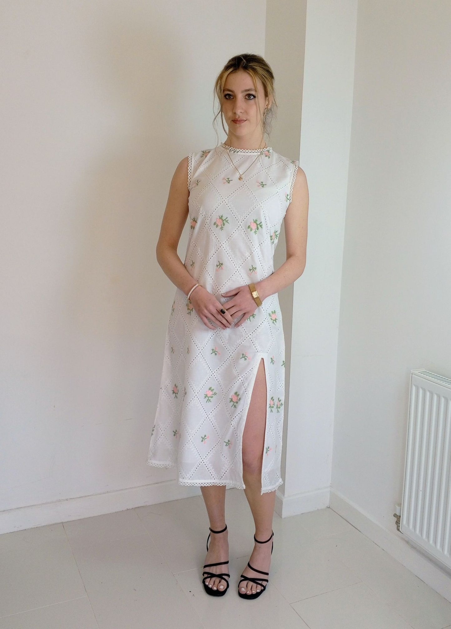 Woman wearing a white floral dress in a minimal room.Pink and white embroidered dress-Midi length relaxed body fit-Boat neck with lace -left side leg split-full lined dress with soft 100% fine cotton -Sleeveless with lace -Fabric: 100% Cotton